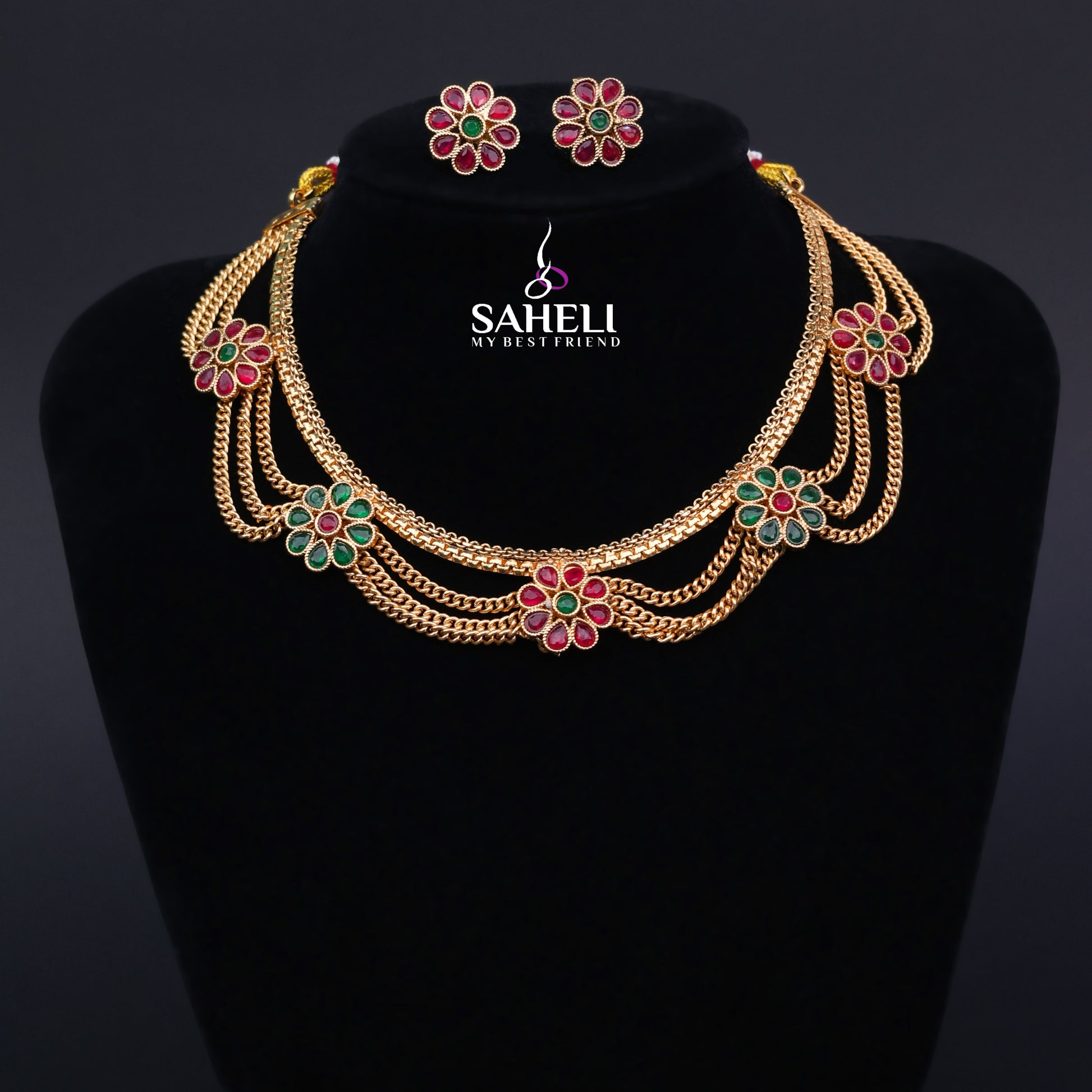 Gold necklace with floral designs and matching earrings on a black stand with 'SAHELI' branding.